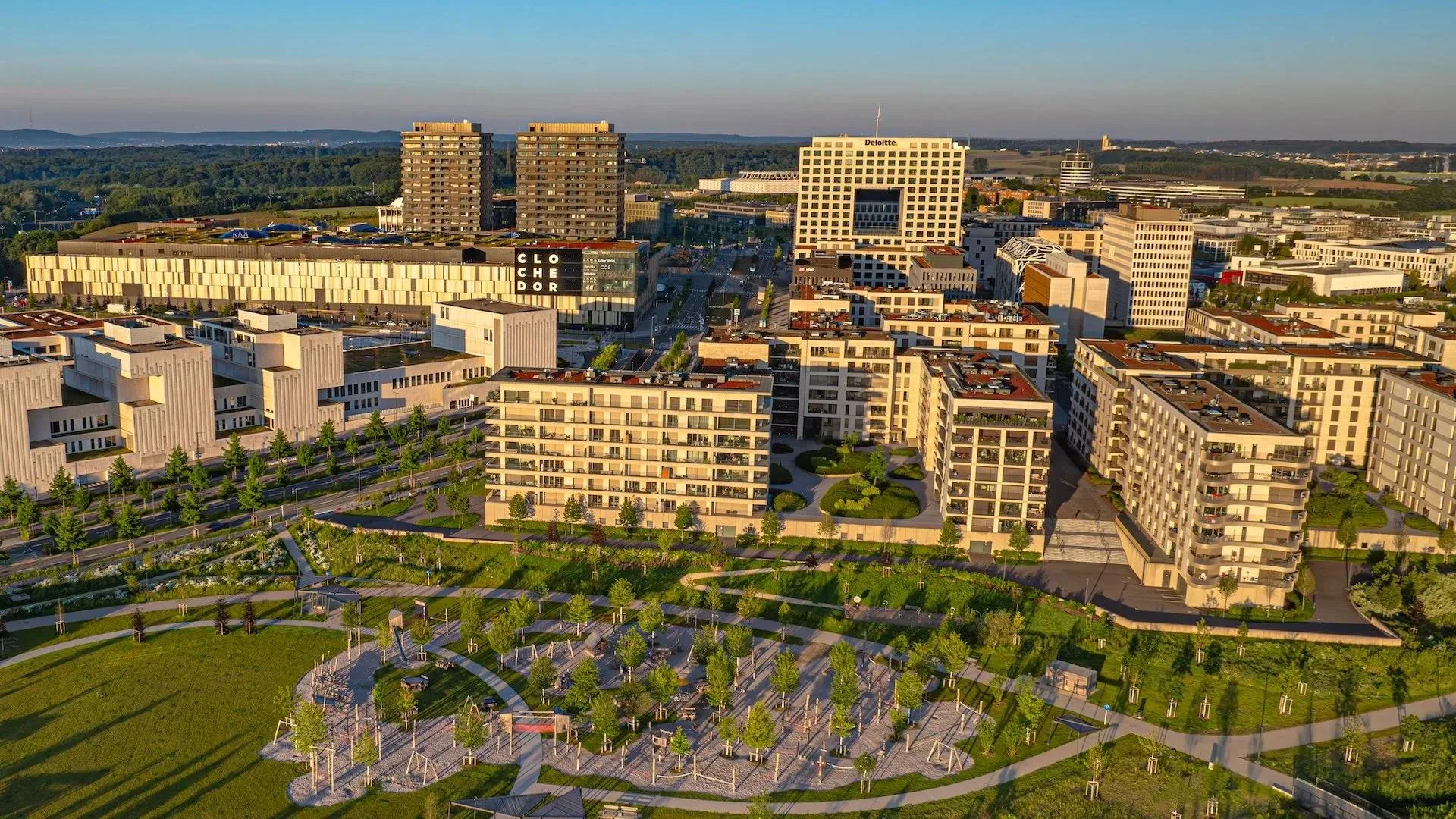Vue aérienne du Parc de Gasperich, ses allées et ses aires de jeux, avec le quartier de la Cloche d’Or en arrière-plan, en lumière de fin de journée